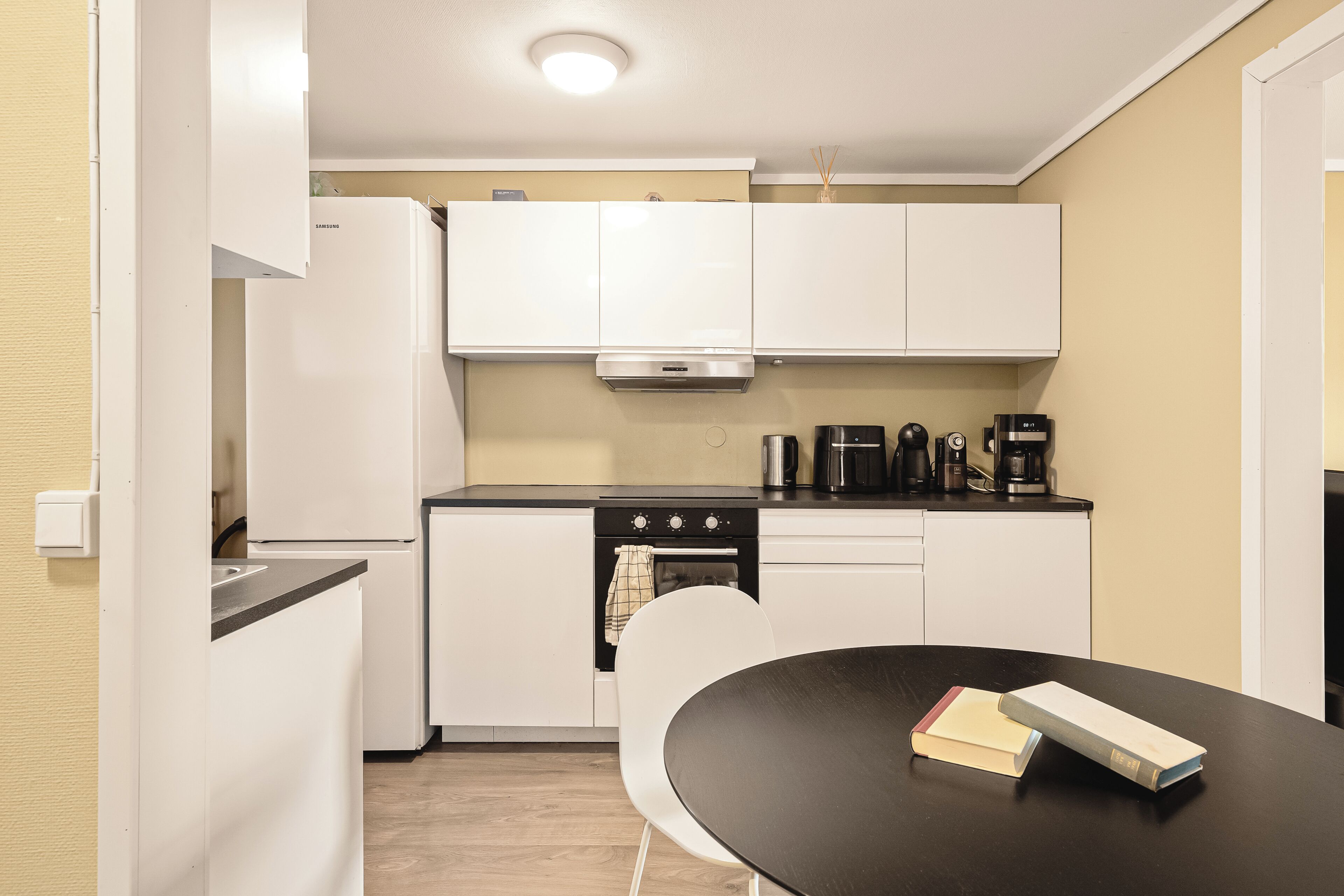 Una cocina compacta con armarios blancos, encimera negra, extractor de acero inoxidable, cafetera y una mesa con libros.