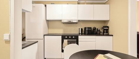 Una cocina compacta con armarios blancos, encimera negra, extractor de acero inoxidable, cafetera y una mesa con libros.