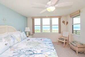 A bedroom with a bed, a chair, a window with a view of the ocean, and a ceiling fan. - Barefoot 217-Second Floor Direct Oceanfront Condo (St. Augustine)