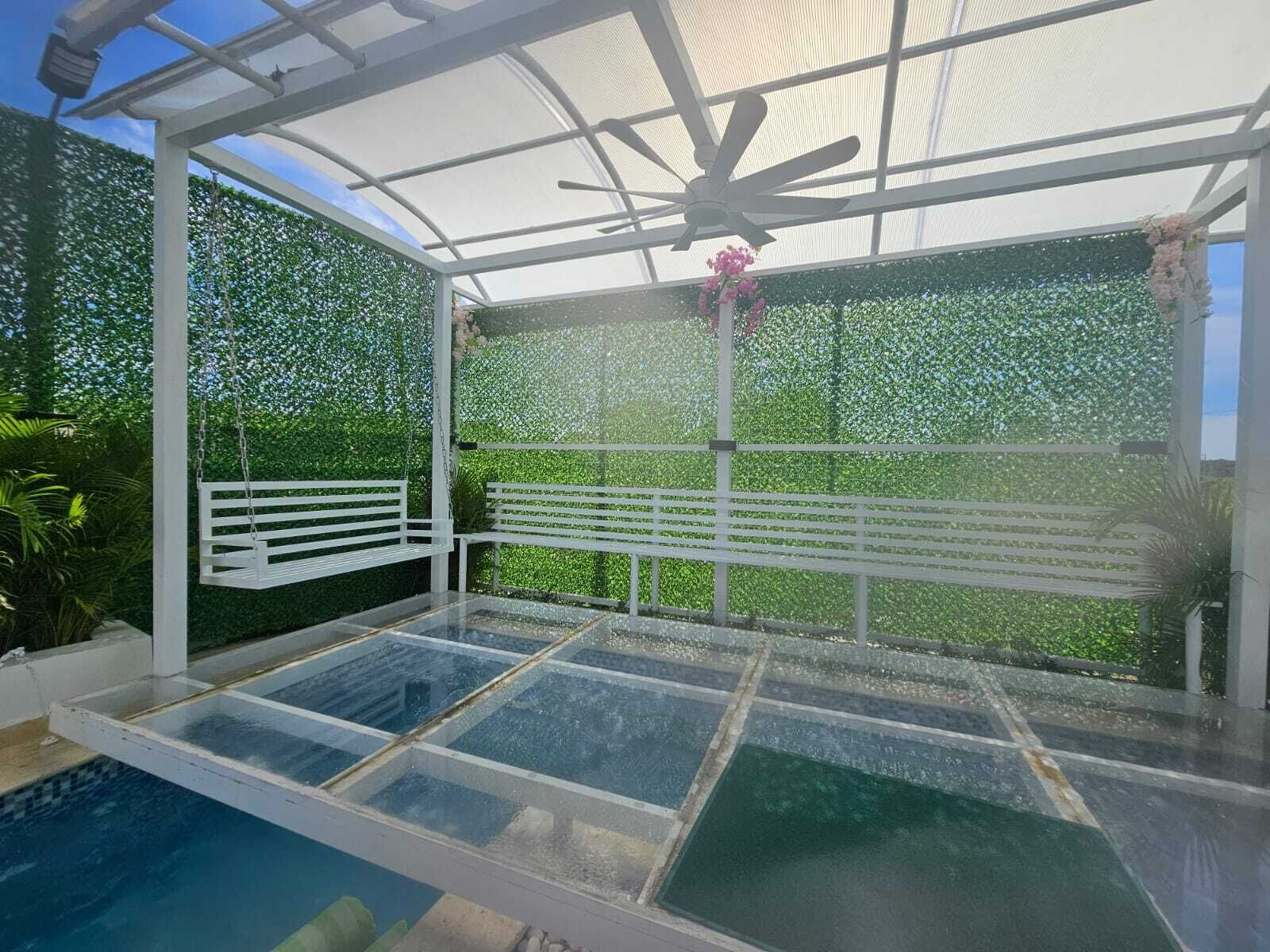 Una piscina exterior cubierta con suelo de cristal, rodeada de vegetación y una valla blanca.