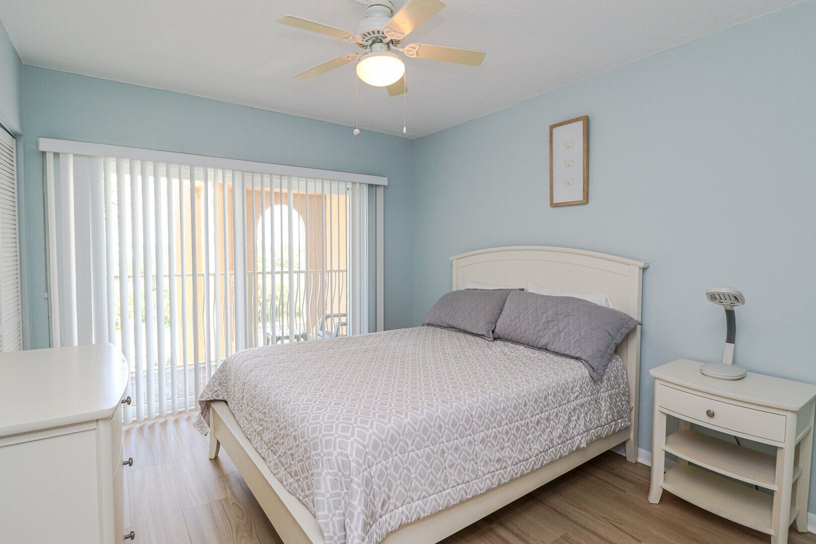 Une chambre à coucher avec un lit, une table de chevet, un ventilateur de plafond et une fenêtre avec des stores.