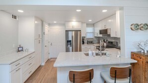 A modern kitchen with white cabinetry, a central island, stainless steel appliances, and a wooden chair. - Island South 2-Beautiful Oceanfront Ground Floor, Two Bedroom Condo (St. Augustine)