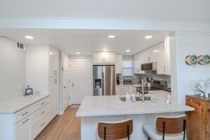A modern kitchen with white cabinetry, a central island, stainless steel appliances, and a wooden chair. - Island South 2-Beautiful Oceanfront Ground Floor, Two Bedroom Condo (St. Augustine)