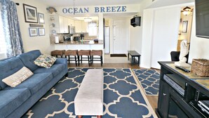 A living room with a blue sofa, a coffee table, and a patterned rug. There is a kitchen in the background with a dining area. - Ocean Village Club Condo D21 (St. Augustine)