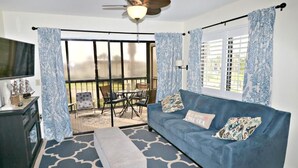 A living room with a blue sofa, a television, a ceiling fan, and a balcony with a table and chairs. - Ocean Village Club Condo D21 (St. Augustine)