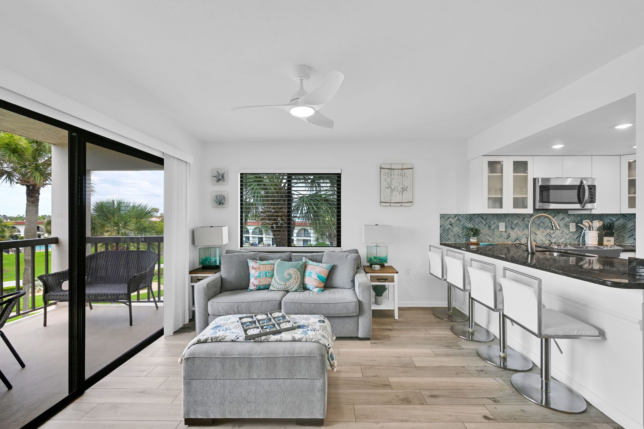 Un interior moderno con un sofá gris, una zona de comedor con taburetes de bar, una cocina con salpicadero en espiga y un balcón con mobiliario de exterior.