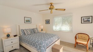 Ein Schlafzimmer mit Bett, Nachttischen, einem Sessel, einem Deckenventilator und einem Fenster mit Jalousien.