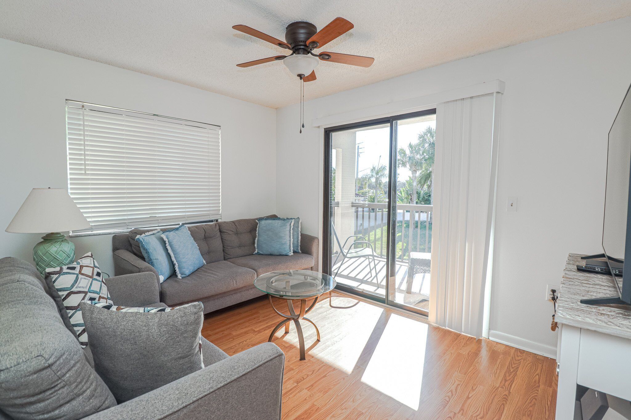 A living room with a sofa, a glass coffee table, a ceiling fan, and a view of the outdoors through a sliding glass door.