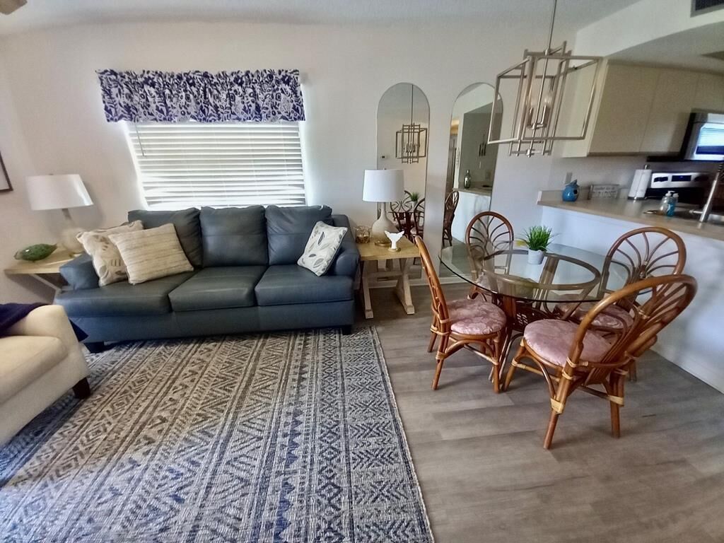 A living room with a sofa, a dining table with chairs, a patterned rug, and a kitchen area in the background.