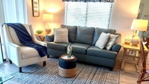 Una sala de estar con un sofá gris, un sillón azul, una mesa redonda de centro y una alfombra estampada.