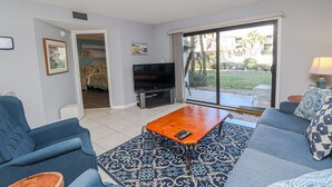 Ein Wohnzimmer mit Fernseher, blauen Sofas, einem Couchtisch und Gartenblick durch eine Schiebetür.