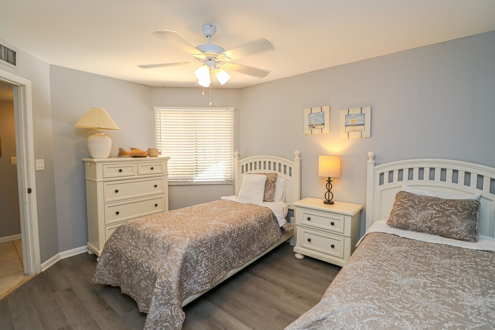 A bedroom with two beds, a ceiling fan, a window with blinds, a nightstand with a lamp, and a dresser.