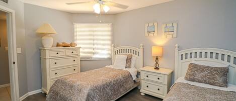 A bedroom with two beds, a ceiling fan, a window with blinds, a nightstand with a lamp, and a dresser.