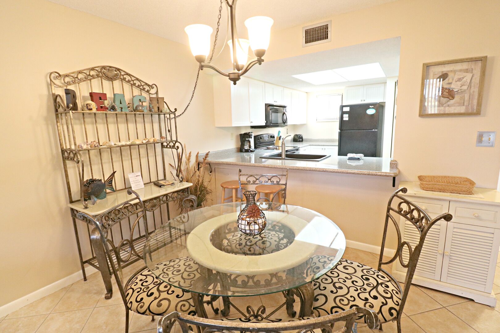 A dining area with a round glass table, metal chairs, a chandelier, and a kitchen in the background.