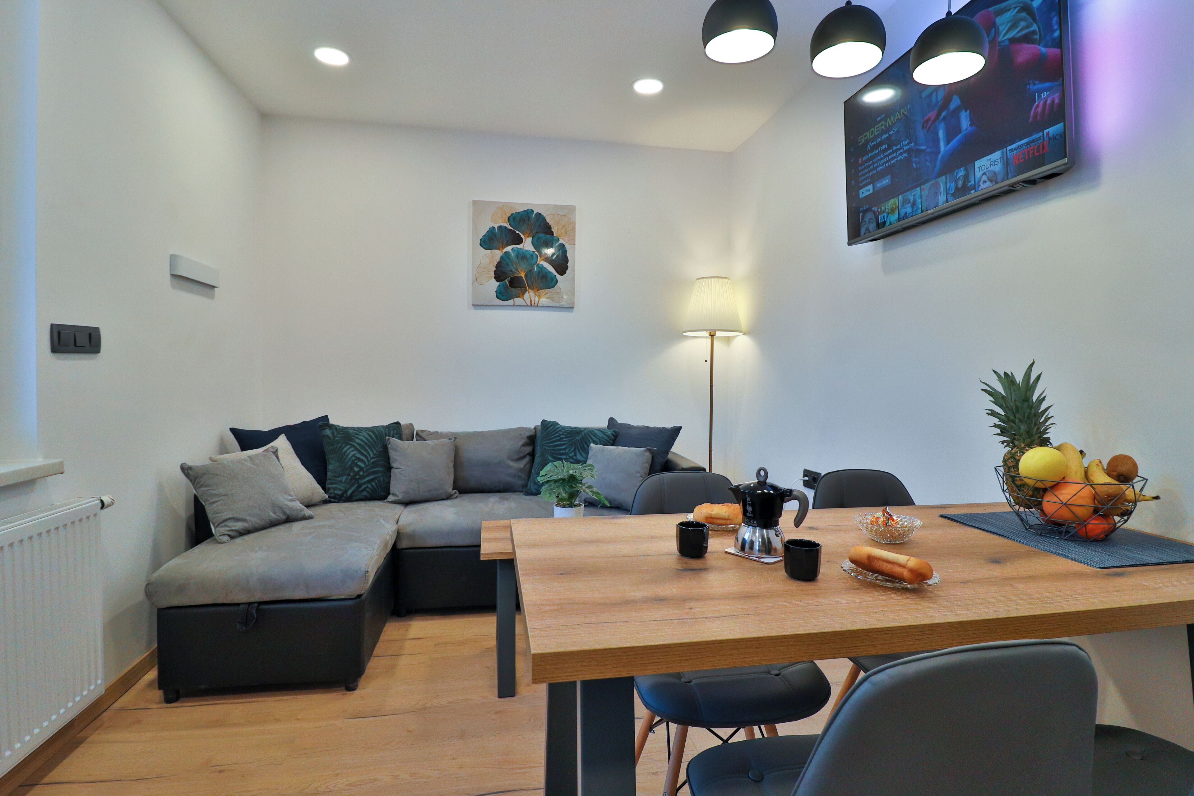A modern living room with a wooden dining table, a sofa, and a flat-screen TV mounted on the wall.