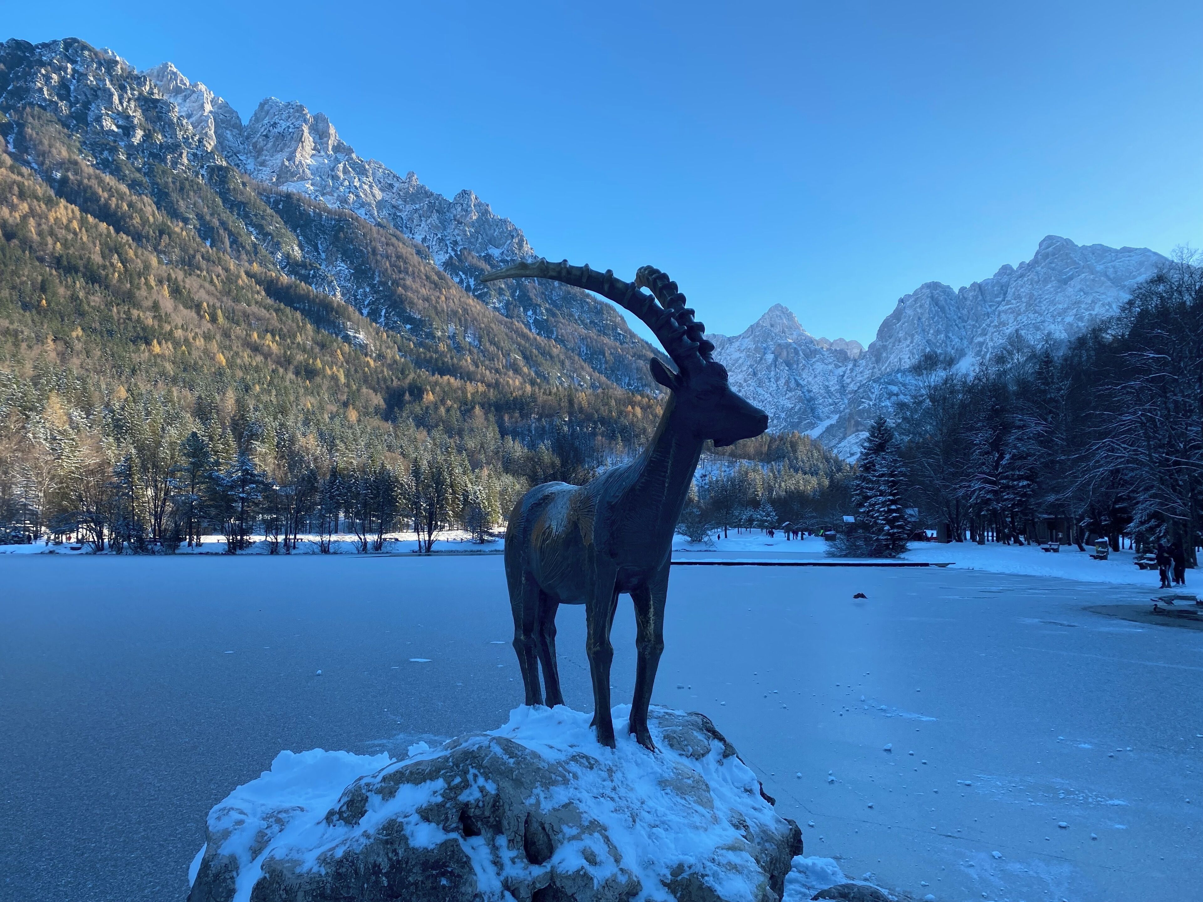 Una estatua de ciervo sobre una roca, junto a un lago con montañas nevadas al fondo.