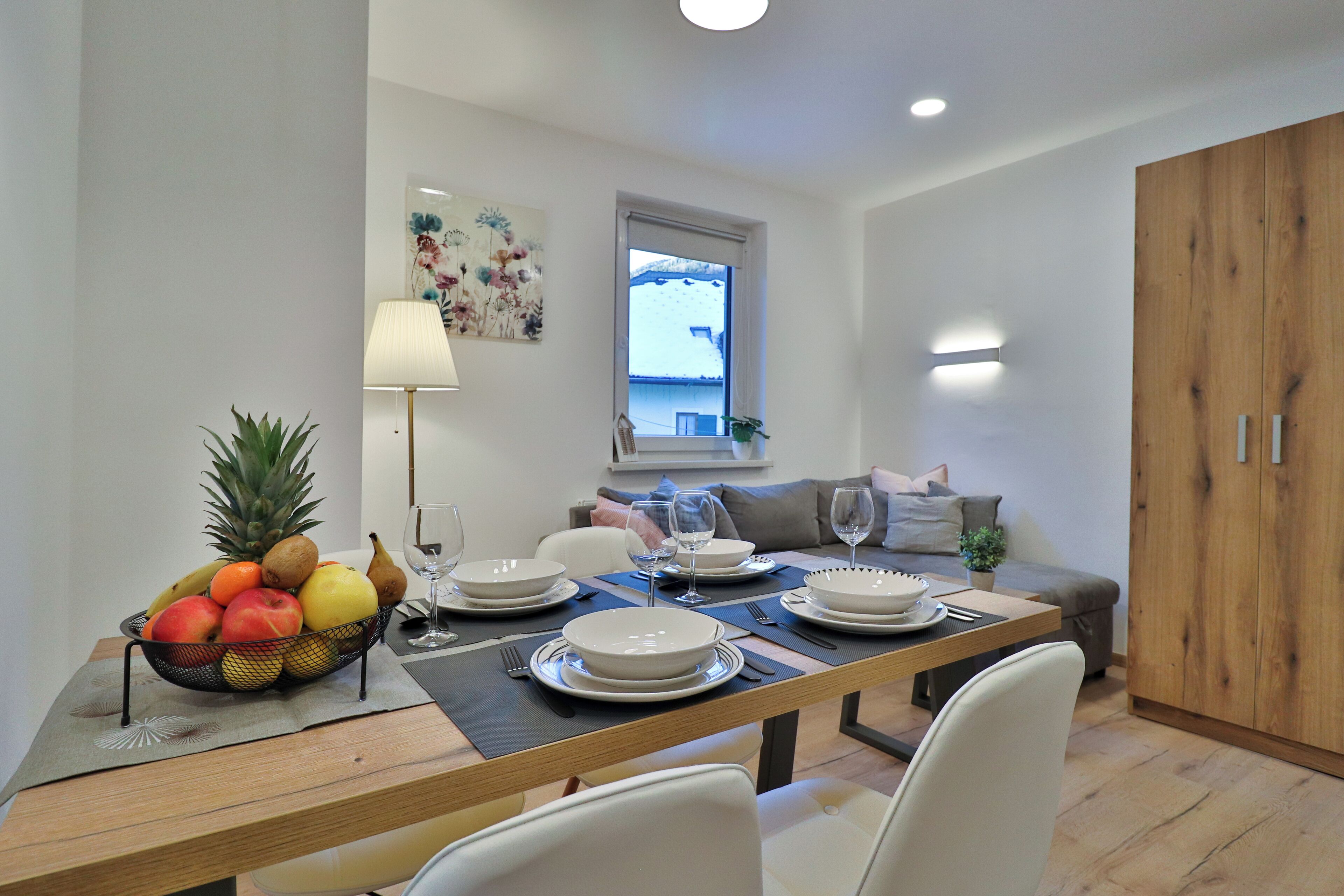 Un comedor con una mesa de madera dispuesta para cuatro personas, rodeada de sillas blancas. Hay un frutero sobre la mesa y una ventana con vistas a una casa.