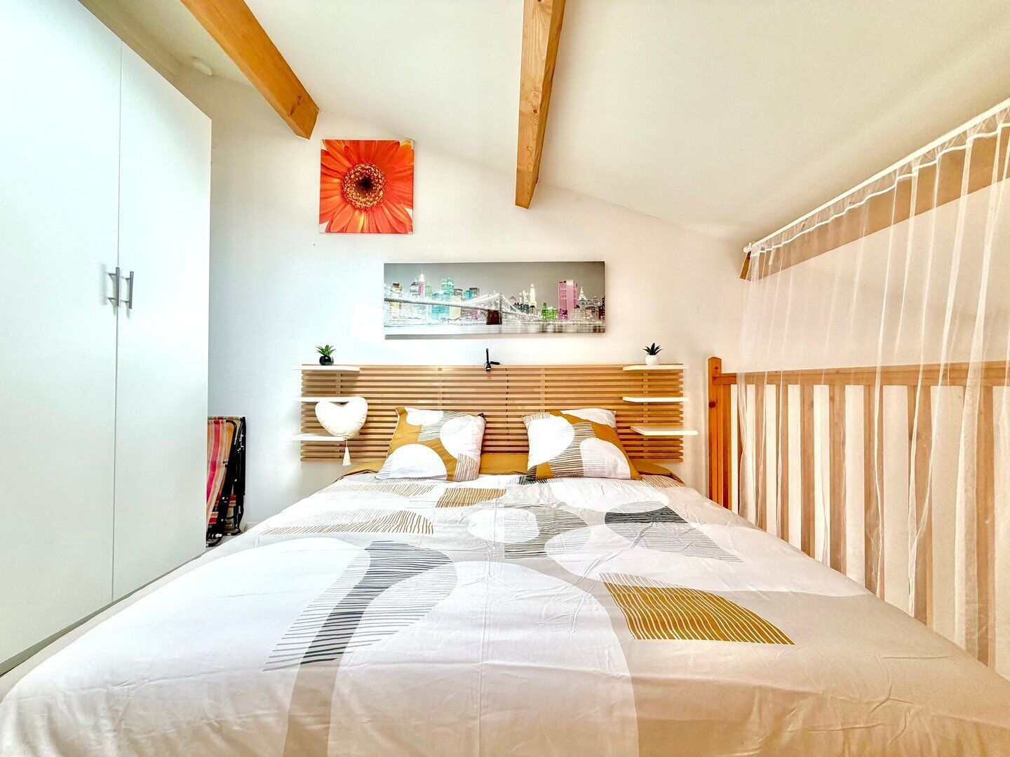 A bedroom with a bed, a cityscape painting, a wooden headboard, and a red flower decoration.