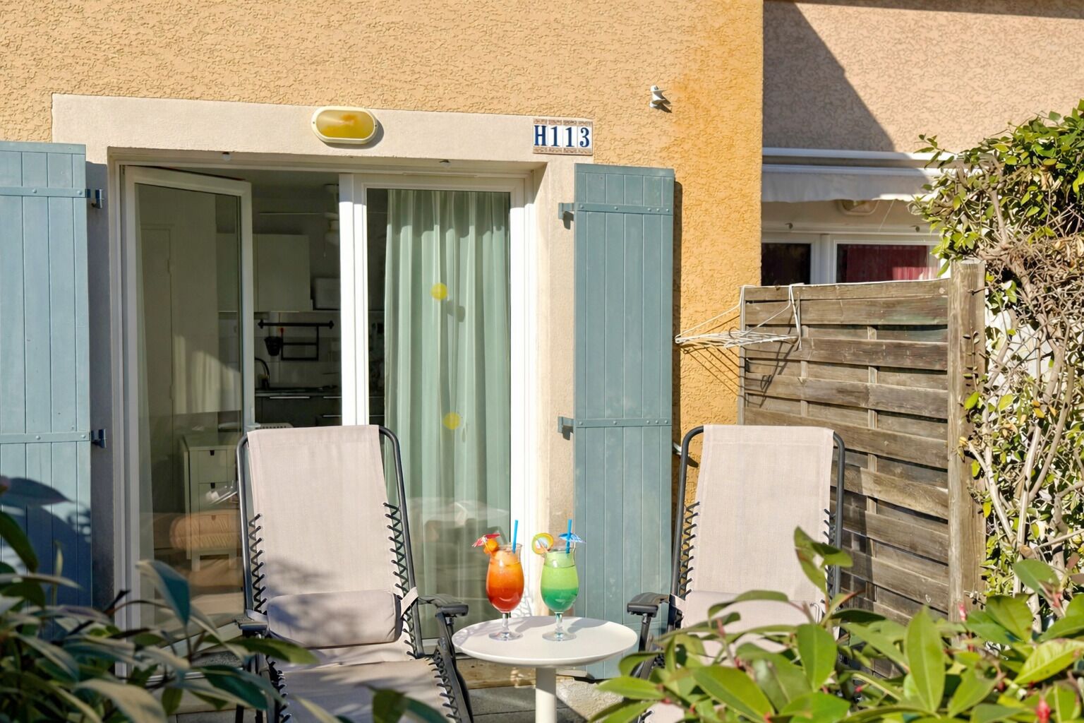 A patio with two chairs and a table, a door with a sign reading "H113", and a small garden area with plants.