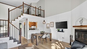 A spacious living area with a wooden staircase, a dining table with chairs, a flat-screen TV mounted on the wall, and a fireplace. - 426 Hotel du Lac (Mont-Tremblant)