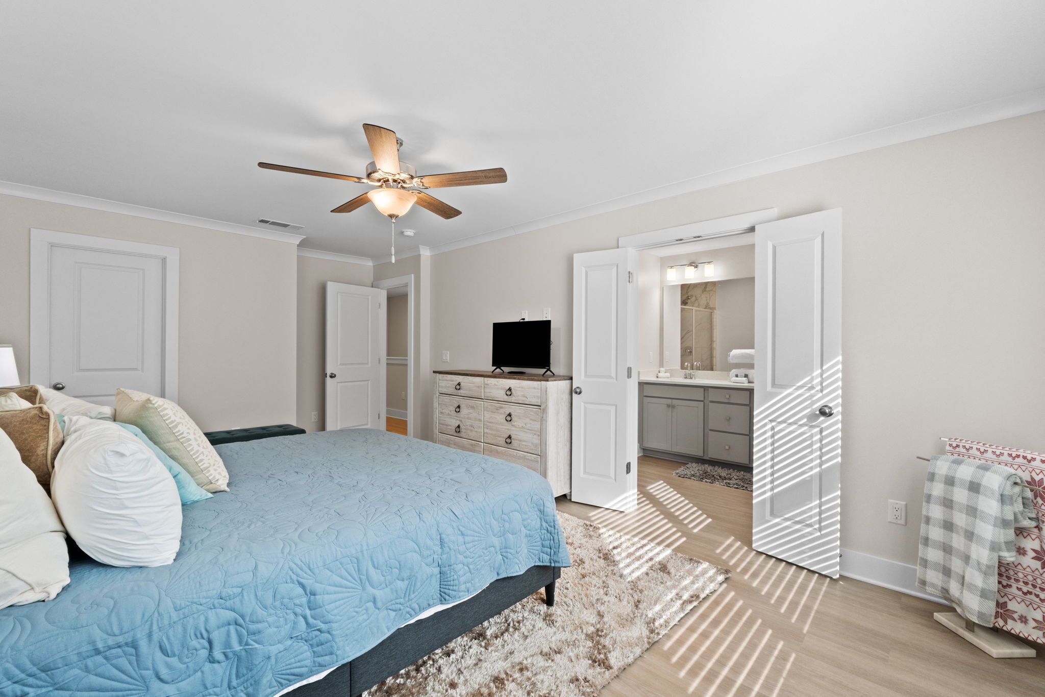 A bedroom with a bed, a ceiling fan, a dresser, and a bathroom visible through an open door.