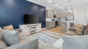 Ein modernes Wohnzimmer mit Flachbildfernseher, grauen Sofas, einem Couchtisch und einer Küchenzeile mit Schränken und Geräten.