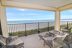 A balcony with wicker chairs and a table, overlooking a beach with a fence and grassy area. - Sand Dollar 1-506-Four Bedrooms, Direct Oceanfront Condo (St. Augustine)