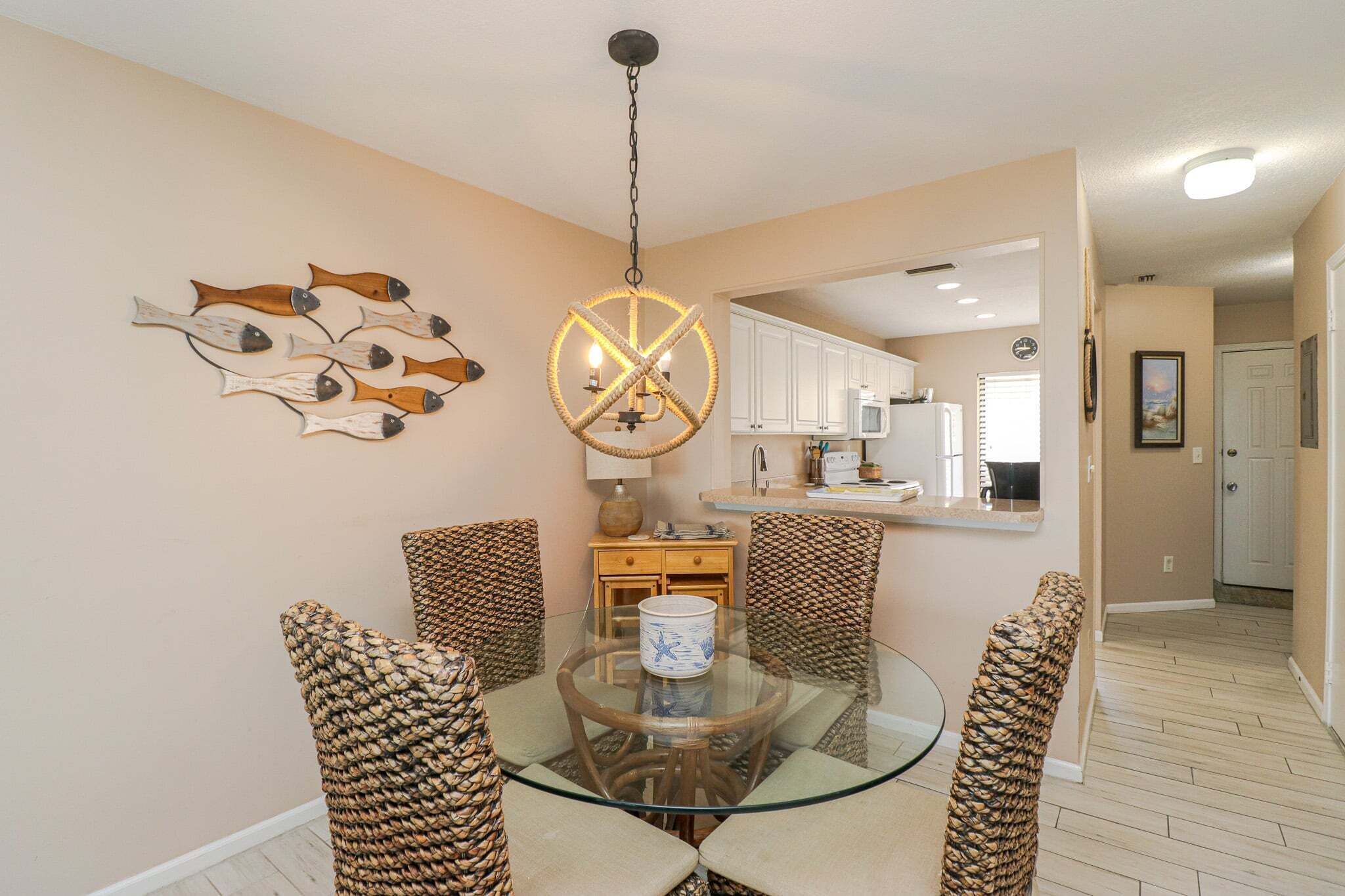 A dining area with a glass table, woven chairs, a decorative fish wall art, a unique chandelier, and a kitchen in the background.