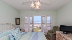 Ein Schlafzimmer mit Bett, Fernseher, Sessel und Blick auf den Strand durch geöffnete Fensterläden.
