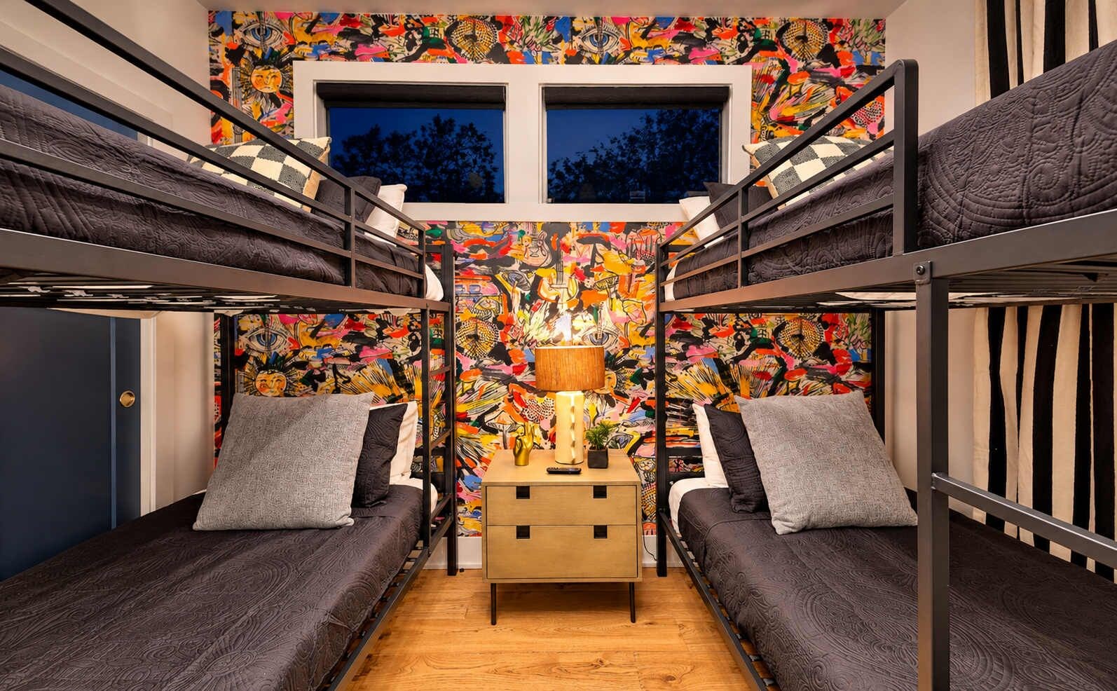A bunk bed room with a colorful mural, a bedside table with a lamp, and a window above the beds.