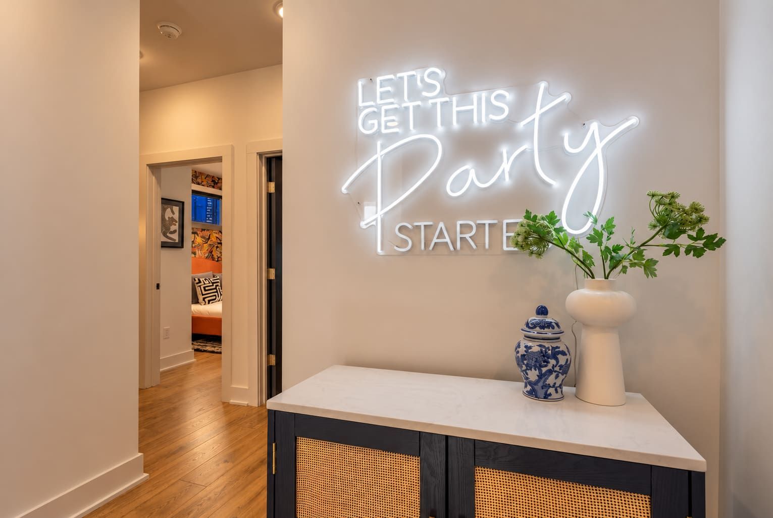 Un pasillo con un letrero de neón que dice 'LET'S GET THIS PARTY STARTED' y un jarrón decorativo con follaje.