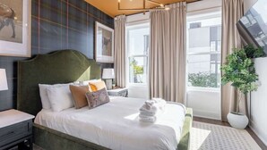 A hotel room with a large bed, a green headboard, a nightstand, a lamp, a framed picture on the wall, a window with curtains, and a potted plant. - 2 Music City Homes | 20 Beds | Firepit & Hot Tub (Nashville)