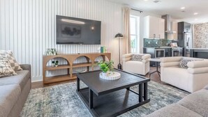 Ein modernes Wohnzimmer mit Flachbildfernseher, zwei Sofas, einem Couchtisch und einer Küchenzeile mit Schränken und Geräten.