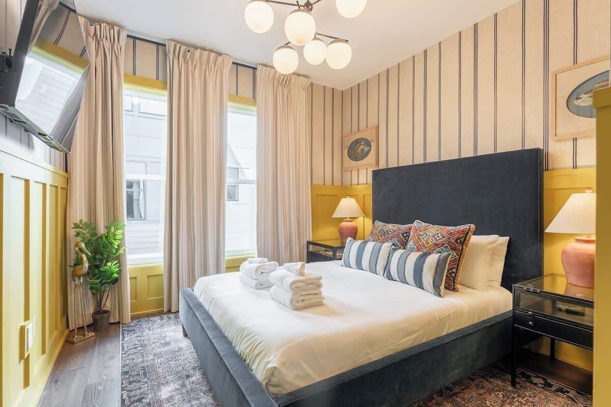 A modern hotel room with a large bed, a small bedside table, a potted plant, and a view of the city.