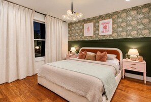 Ein Schlafzimmer mit einem großen Bett, zwei Nachttischen, einem Fenster mit Vorhängen, einer Wand mit gerahmten Bildern und einer Hängelampe.