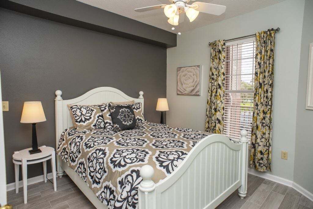 A bedroom with a white bed, patterned bedding, two bedside lamps, a ceiling fan, and a window with floral curtains.