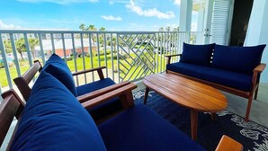 A balcony with a wooden table and two blue cushioned chairs, overlooking a well-maintained lawn and buildings. - Seaside at Anastasia G302- Luxury at the Beach! Pool, Wifi, Elevator, Parking (St. Augustine)