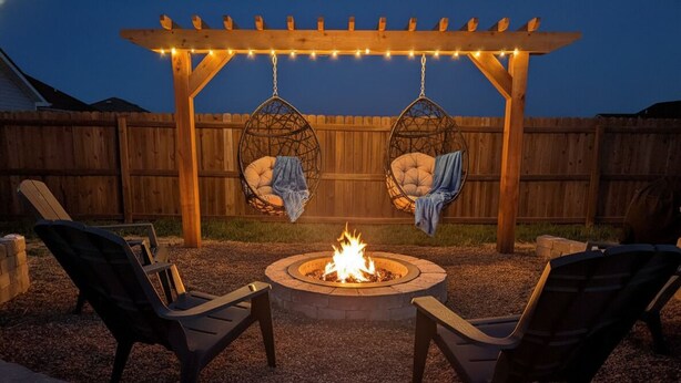 Un foyer extérieur avec deux chaises et deux fauteuils suspendus, le tout installé sous une pergola en bois illuminée de guirlandes lumineuses.