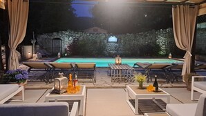 En udendørs terrasse med loungesenge, swimmingpool og en stenvæg med port.