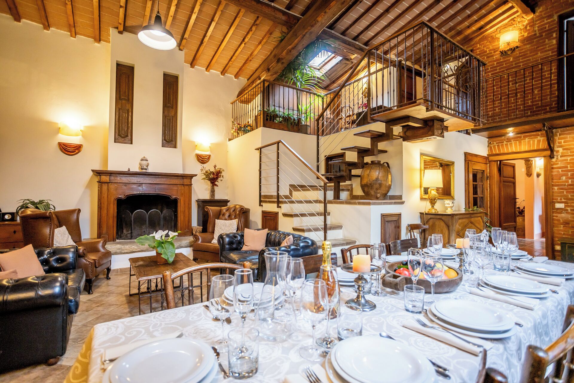 Un comedor con una mesa larga dispuesta para una comida, una chimenea, sillones de cuero y una escalera que sube a un nivel superior.