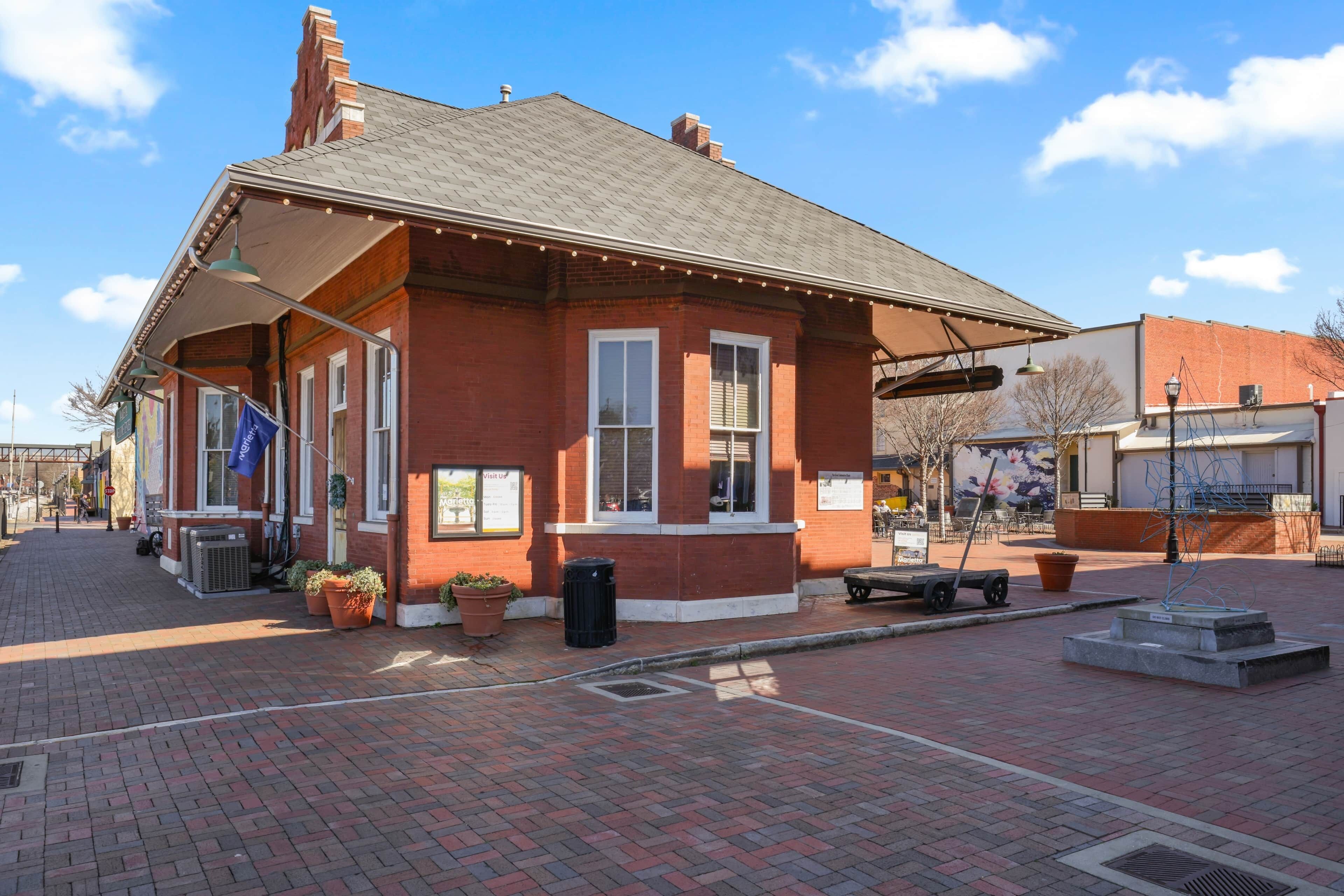 A red brick building with a covered porch, potted plants, and a trash can.
