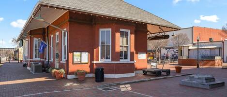 A red brick building with a covered porch, potted plants, and a trash can.