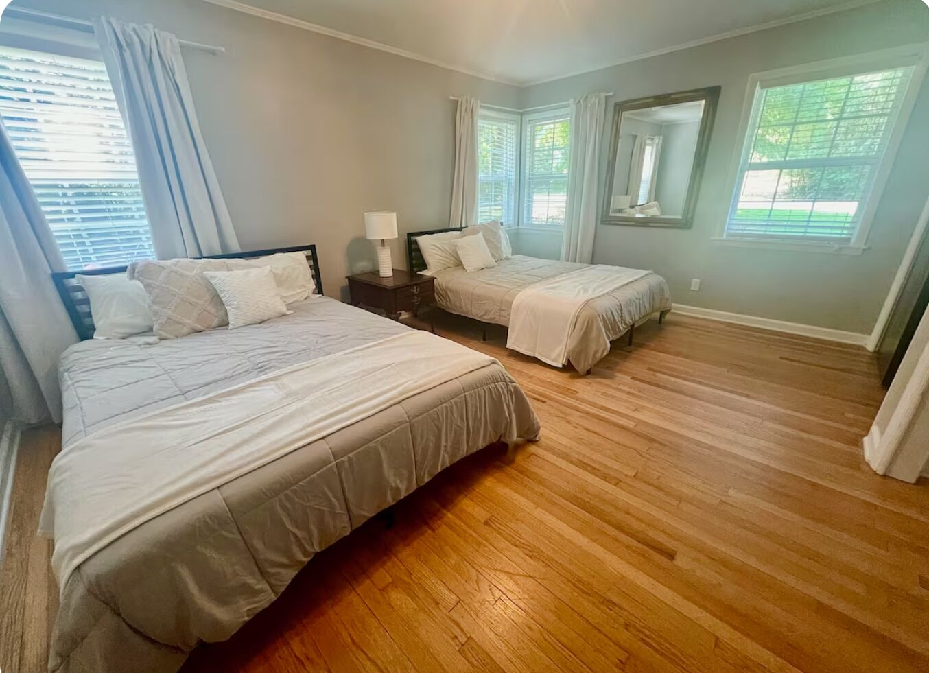 Une chambre avec deux lits, une table de chevet, un miroir et de grandes fenêtres équipées de stores.