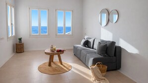 Una sala moderna con un sofá gris, dos espejos redondos, una mesa redonda de madera con una planta y vistas al mar a través de amplios ventanales.