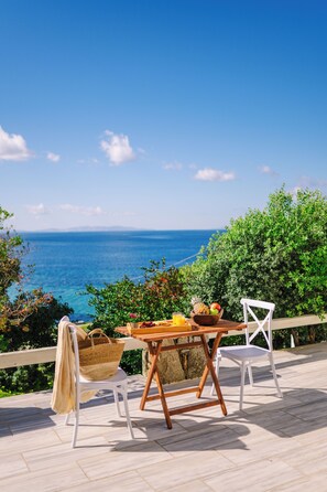 A wooden table with chairs on a deck overlooking the sea, with a basket and fruit on the table.
