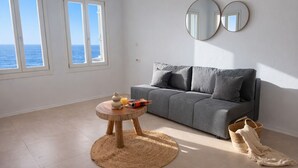 Una habitación con un sofá gris, una mesa redonda de madera con un jarrón y un vaso de jugo de naranja, una alfombra tejida y un ventanal con vistas al mar.