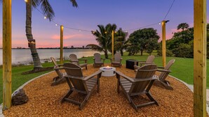 A patio with Adirondack chairs, a fire pit, and string lights against a sunset backdrop. - Waterfront Stunner! Heated Pool+Spa+Sauna+Firepit+ Playground! (West Palm Beach)