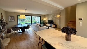 Un vaste espace de vie comprenant une grande table à manger en marbre, un canapé et un téléviseur, le tout adossé à un mur orné d’une fresque représentant une forêt.