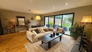 Un salon spacieux avec un grand canapé, une table basse et un coin repas comprenant une table et des chaises. Une grande fenêtre offre une vue sur l’extérieur.