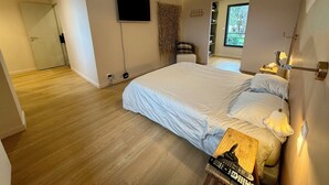 Une chambre avec un grand lit, un téléviseur fixé au mur, une table de chevet avec une lampe et une fenêtre donnant sur un paysage verdoyant.